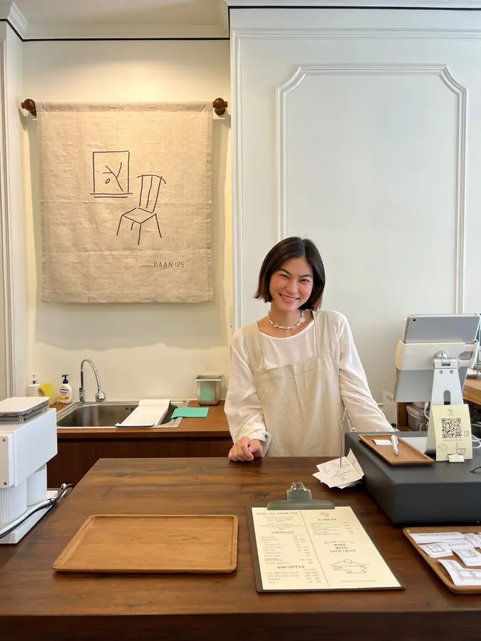 Milk smiling behind the baan125 counter, the BAAN 125 tapestry on the wall behind her and the menu board on the counter