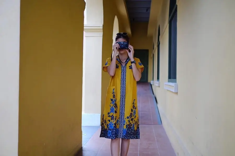Milk wearing a mustard Phathoongnoongdai batik dress with blue florals, holding a film camera in a shophouse corridor